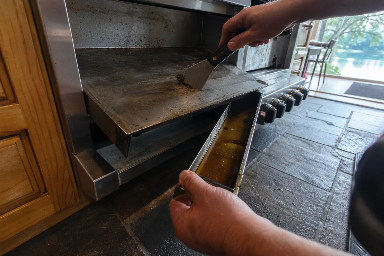 Hand pushing a flat scraper across a stainless range griddle while the other hand holds the pulled-out grease trough, black knobs and slate floor beside it