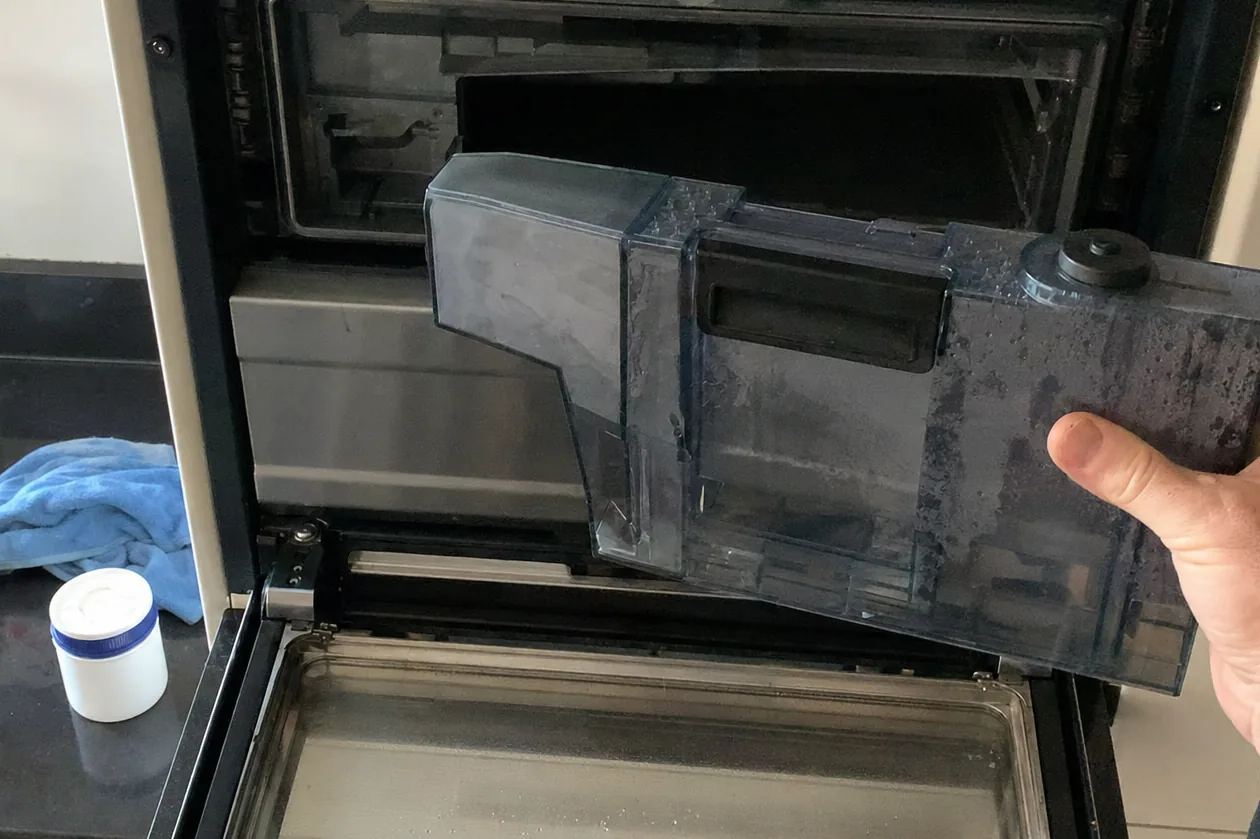 Mirrored close frame of an appliance water tank withdrawn from its slot, scale visible along the seam
